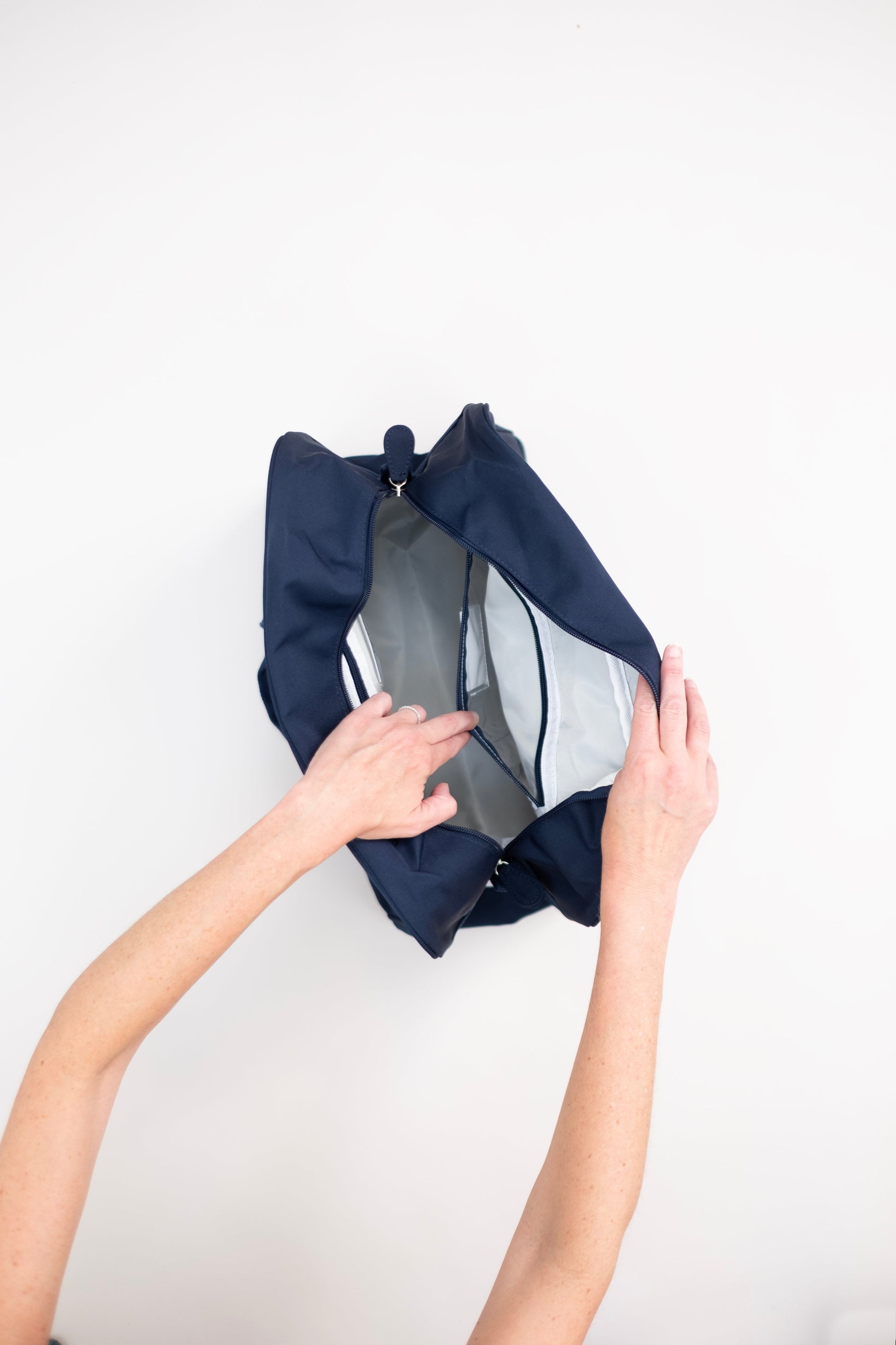Person opening a navy blue duffle bag to show interior pockets with a white interior on a light gray background