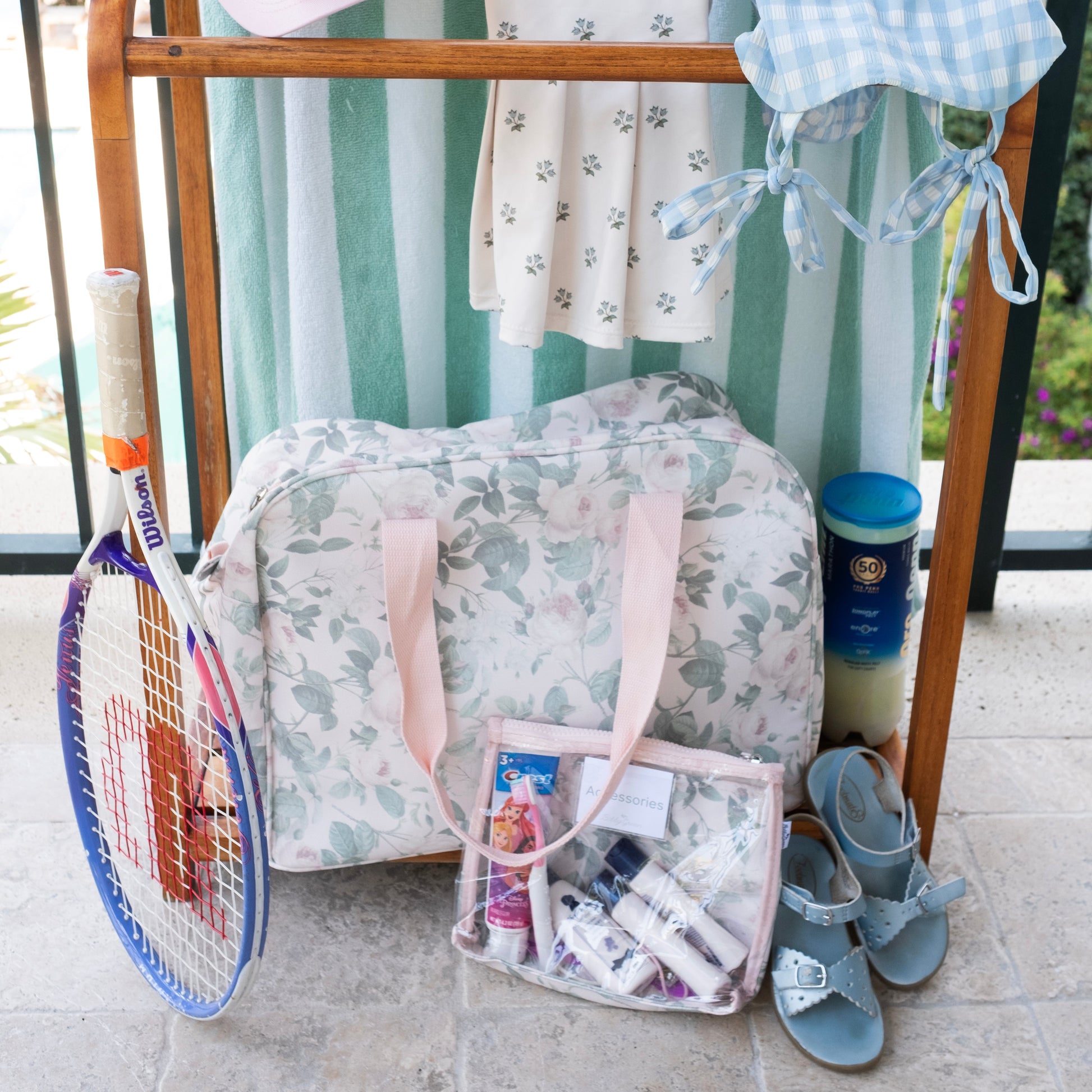 Child travel items including a bag, clothes, and a tennis racket on a wooden rack outdoors.